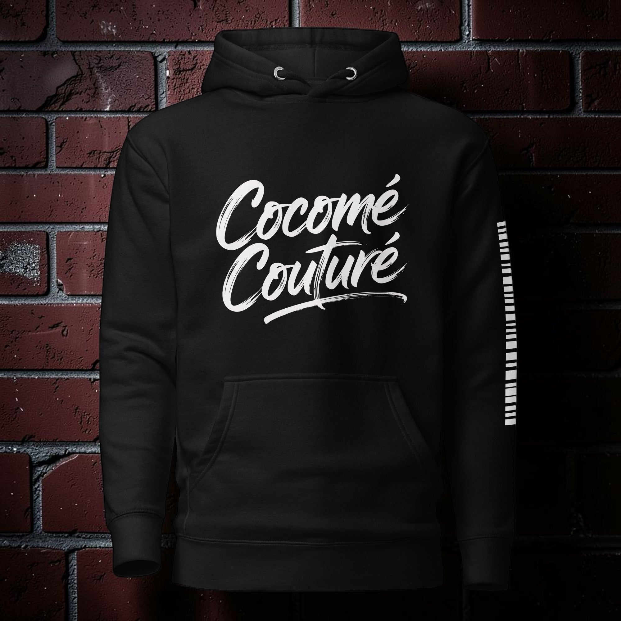 CocoMe Couture black premium hoodie with white brush script logo and kangaroo pocket against a brick wall background